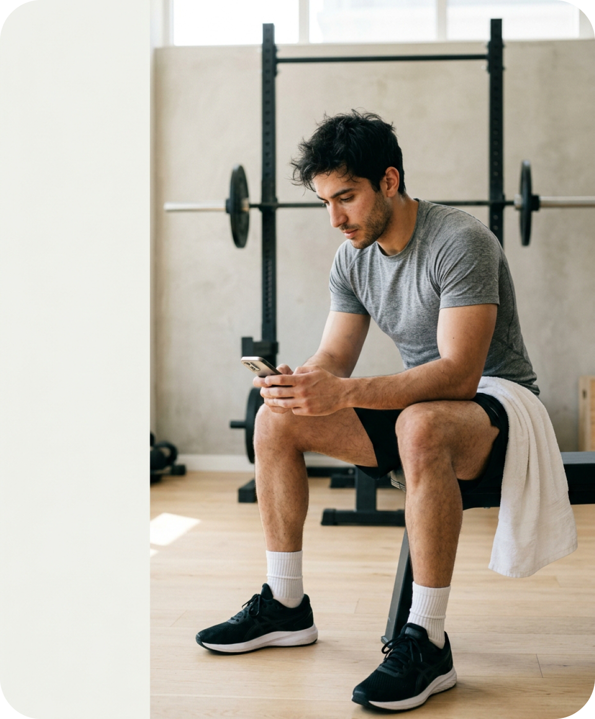 A man sitting on a weight bench in a gym between sets, looking at his phone.
